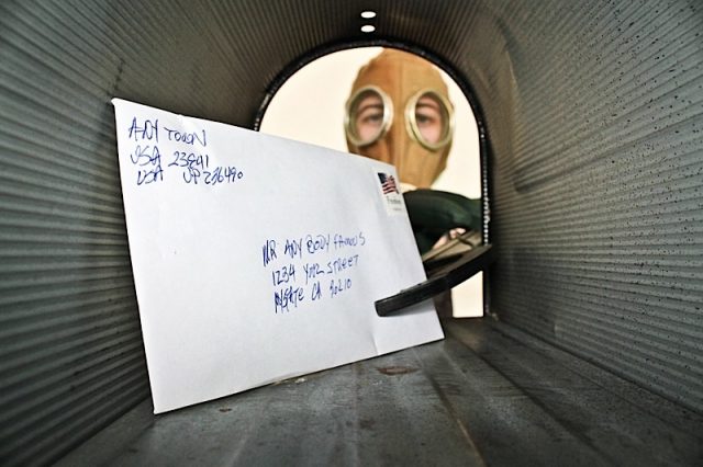A unidentifiable person in a Gas Mask retrieves or sends mail from their mail box with an extension Photo courtesy of BigStock/mikeledray