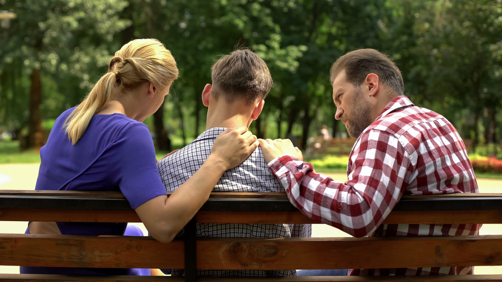 Caring, upset mother and dad supporting their upset son on a park bench.