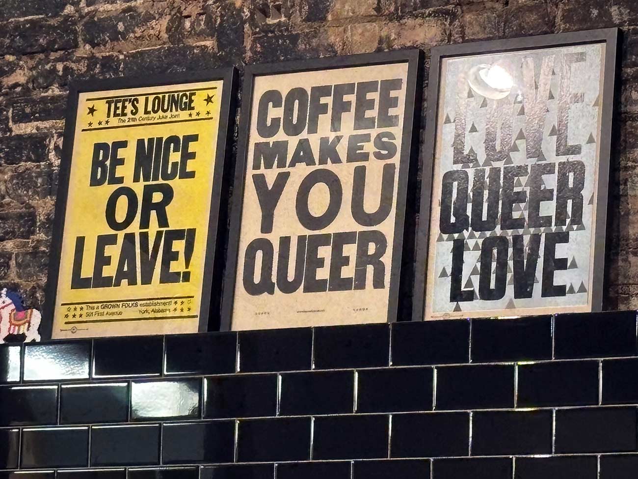 Three signs on a coffee shop wall promoting be nice, coffee, and queer love.