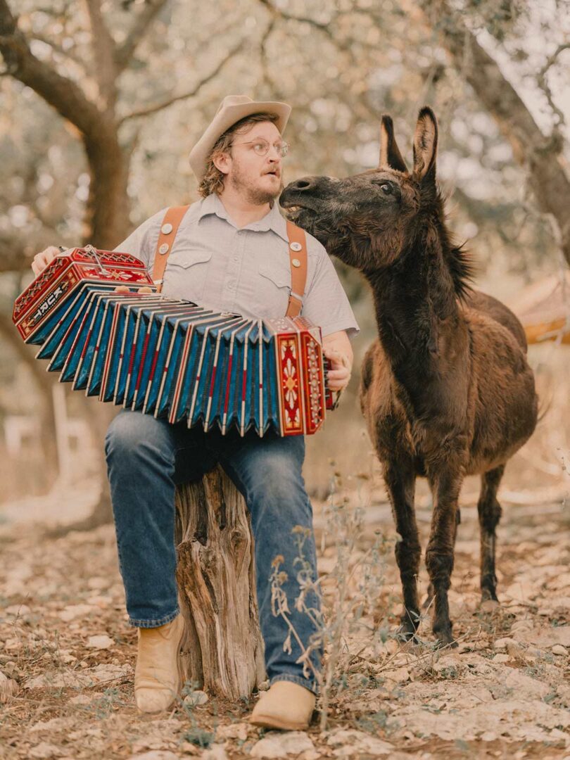 Music artists Willi Carlisle playing in the woods with a donkey standing next to him.