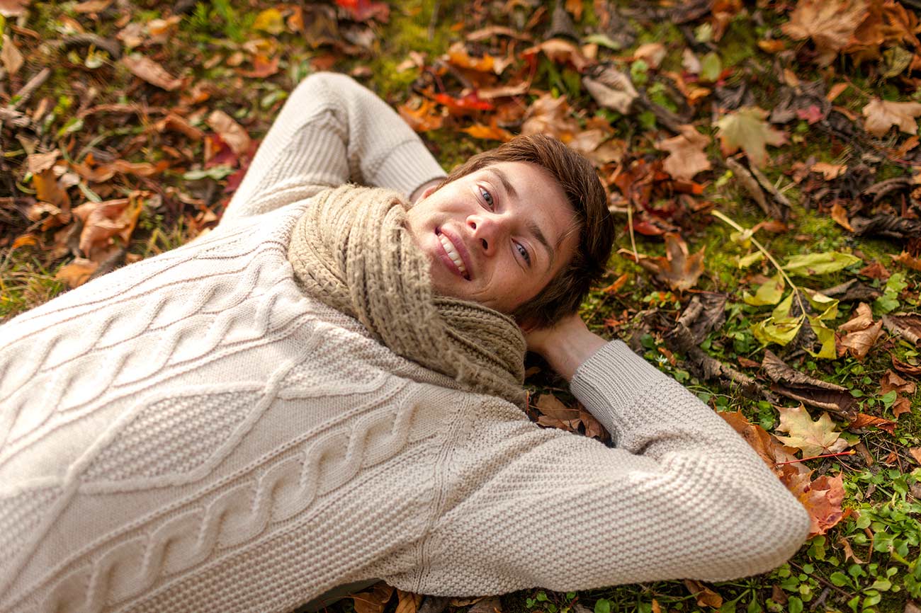 Close up of smiling young man lying on ground or grass and fallen leaves in autumn park.