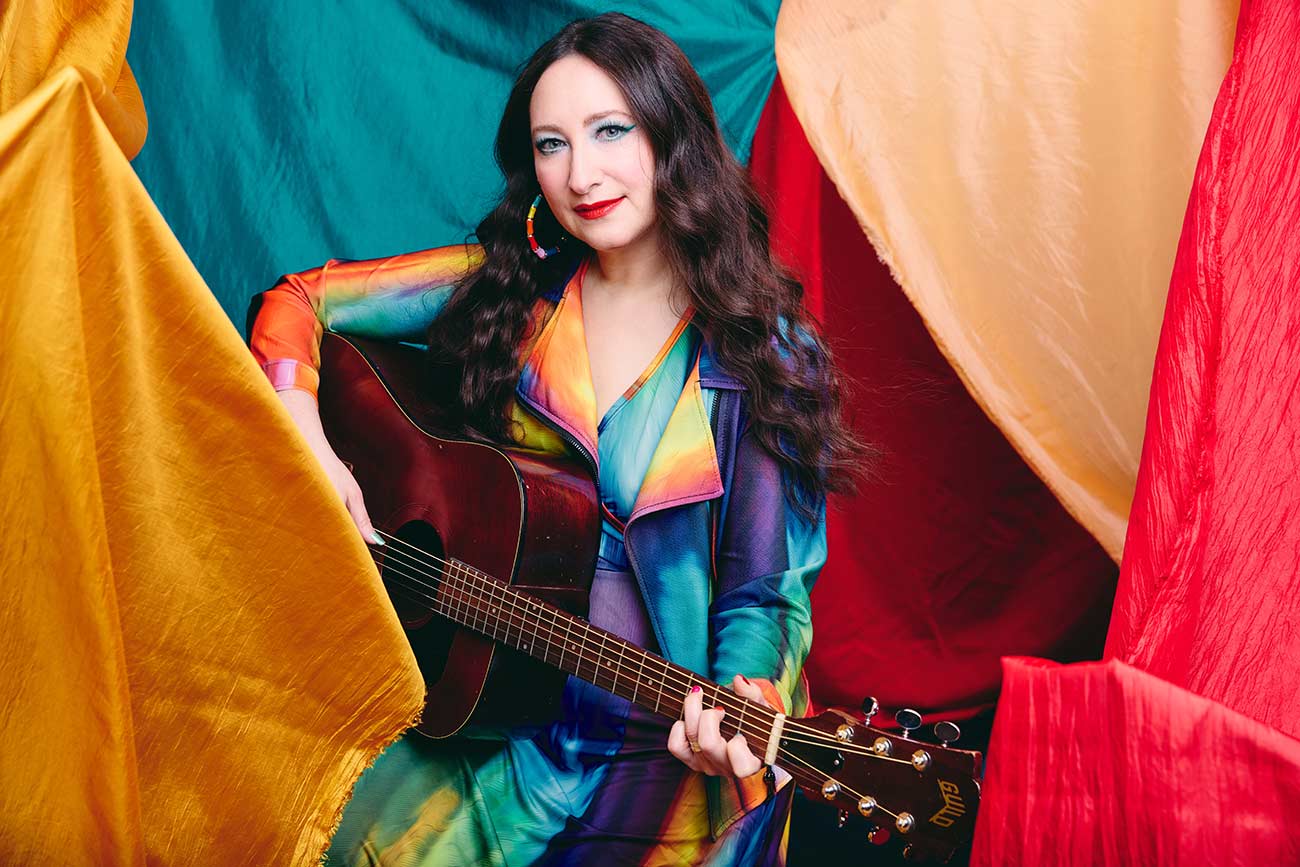 Musician Rachael Sage in a colorful outfit holding her guitar posing for a promotional photo.