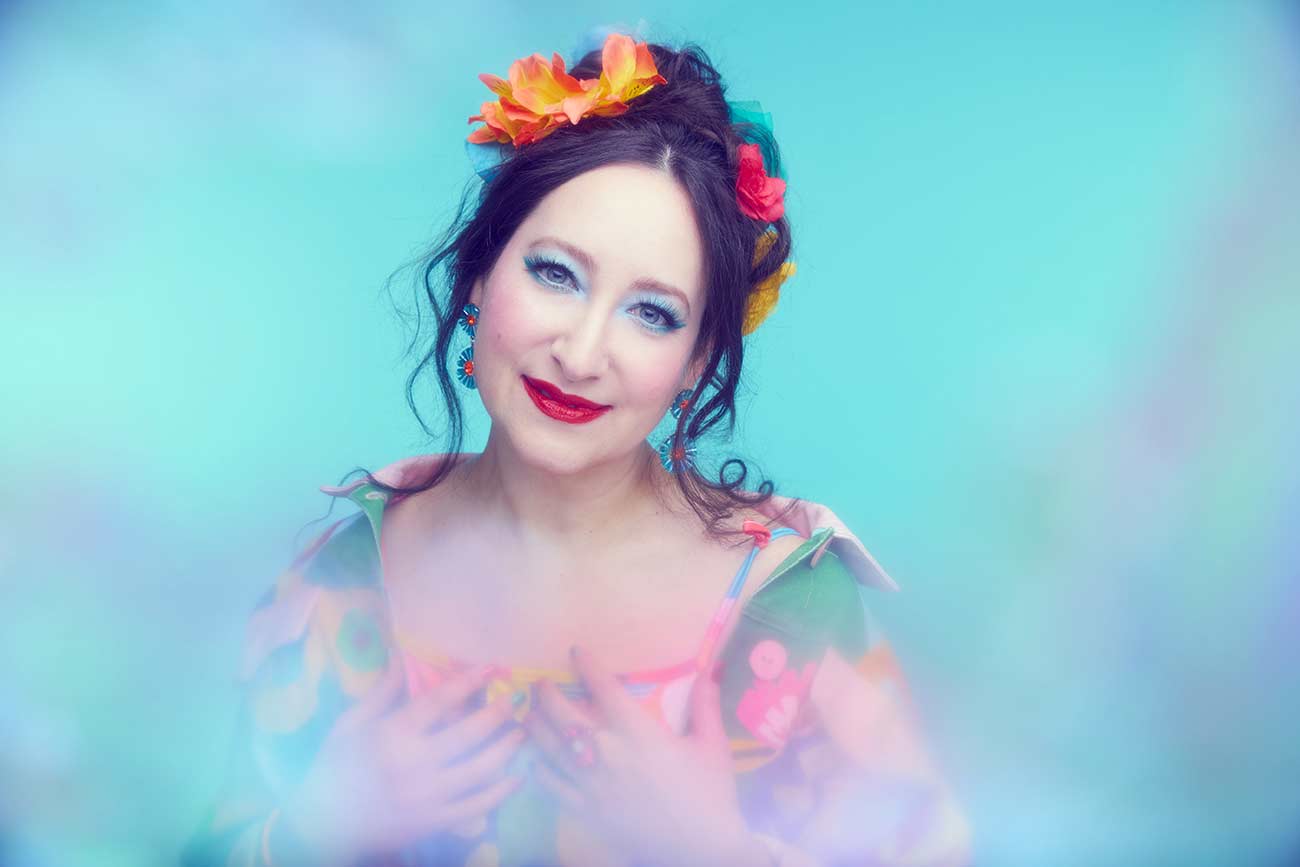 Musician Rachael Sage with colorful flowers in her hair holds her hands to her chest with a soft smile.
