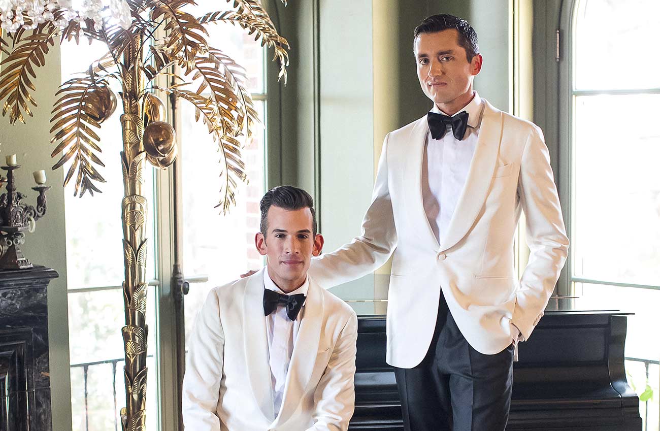 Handsome male couple standing and sitting wearing white tuxedos by a piano.