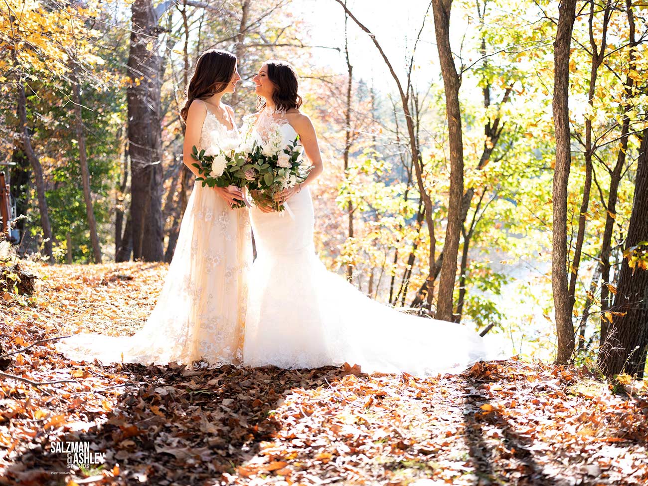 Two young women in white wedding dresses pose together in the fall woods.