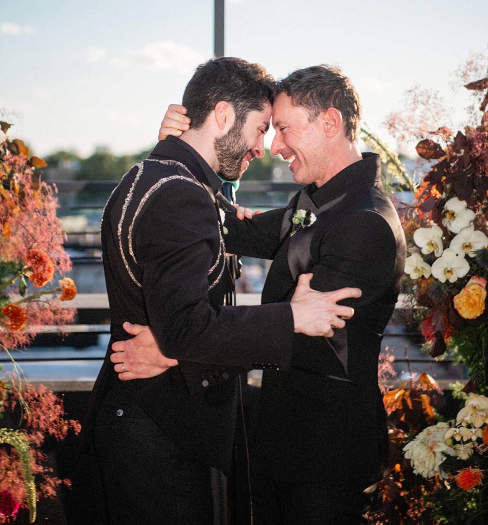 Male couple wearing black tuxedos embrace each other with happy smiles.