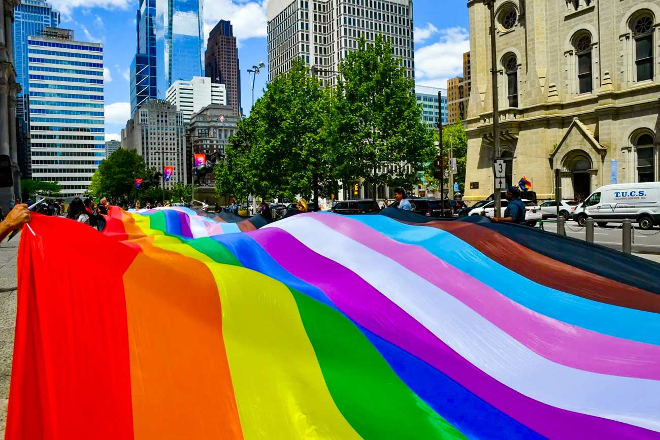 Large rainbow flag flowing in the street during a parade.
