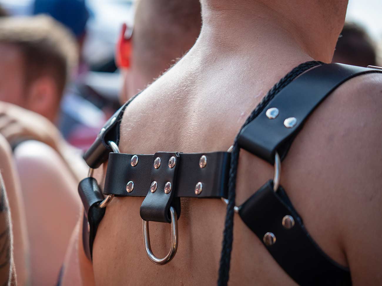 Close up of a leather harness on the back of a shirtless white male in a crowd.
