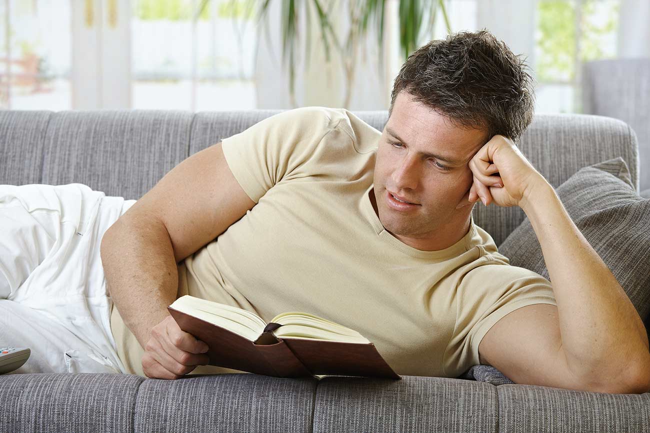 Handsome man in causal wear smiling lying on sofa reading handheld book.