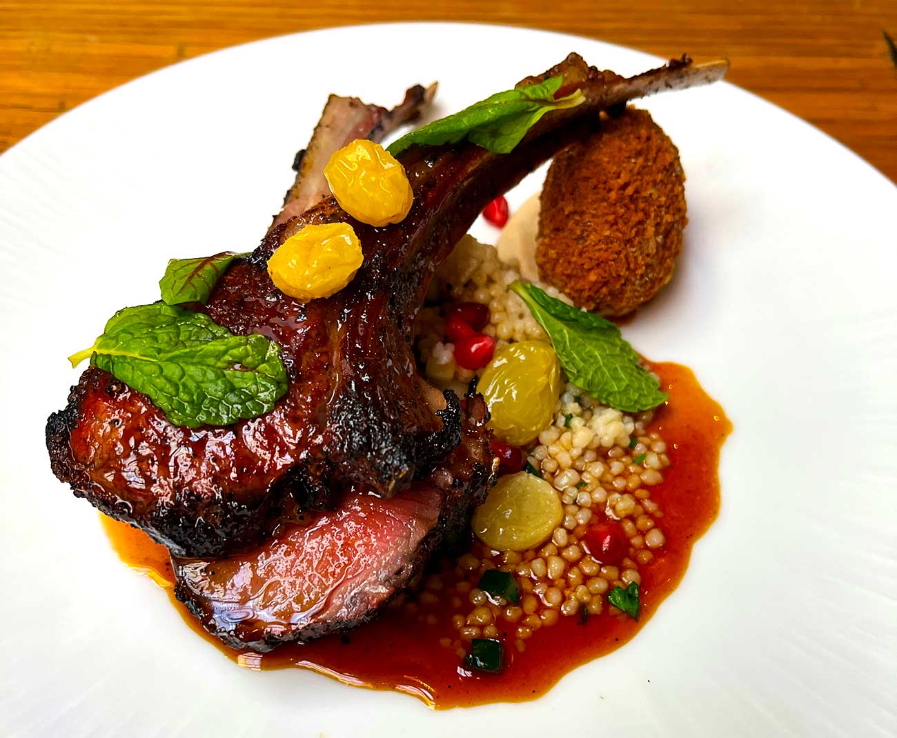 A plate featuring cuts of a rack of lamb on a bed of cous cous with kibbeh and red sauce.