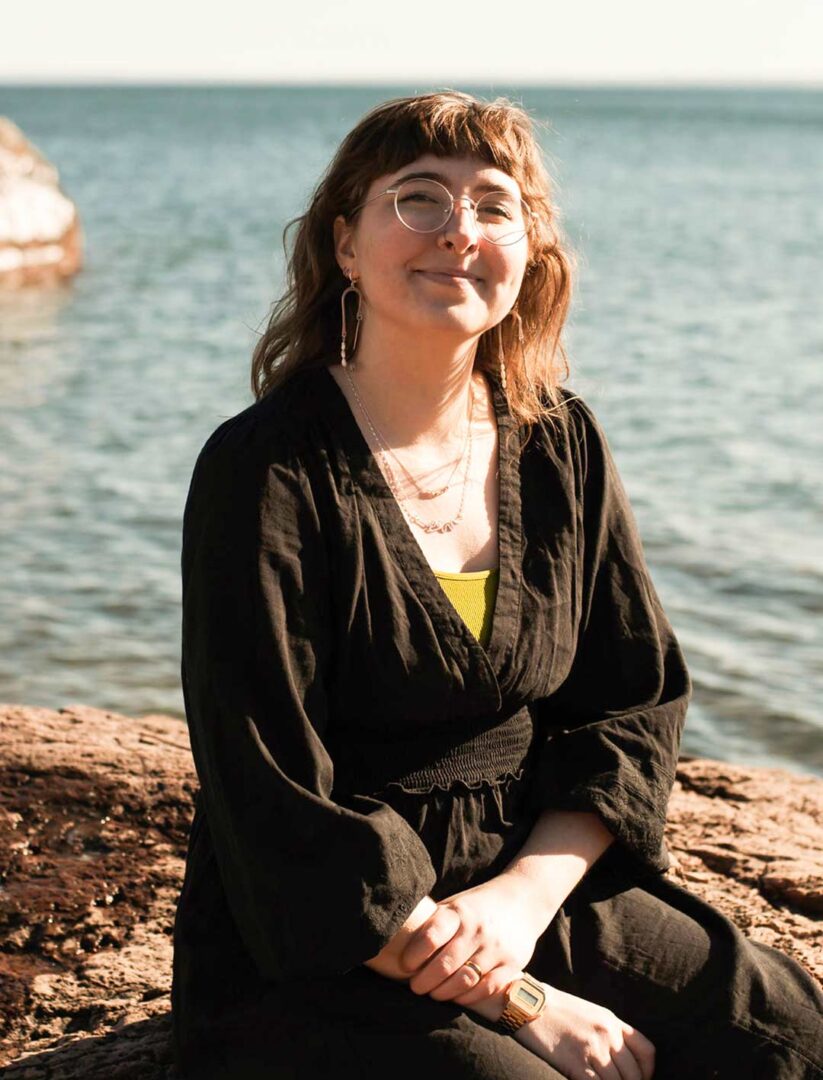 Portrait of Ruby Dammann sitting on the rocks by Lake Superior.