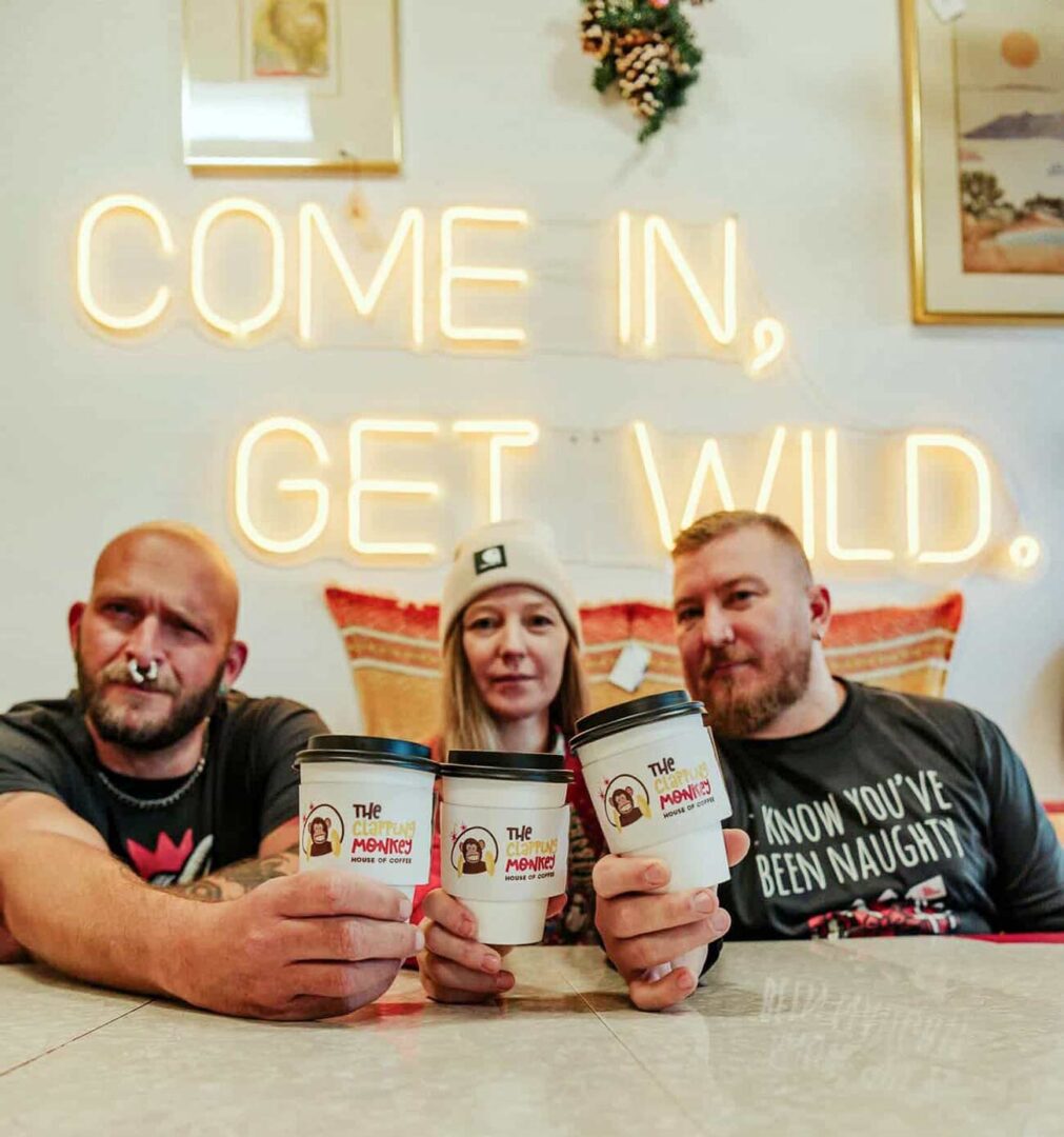 Owner David Wenzel, General Manager of Operations Lindsey Schwendler and Owner Joshua Larson holding their Clapping Monkey coffee cups sitting at a table.