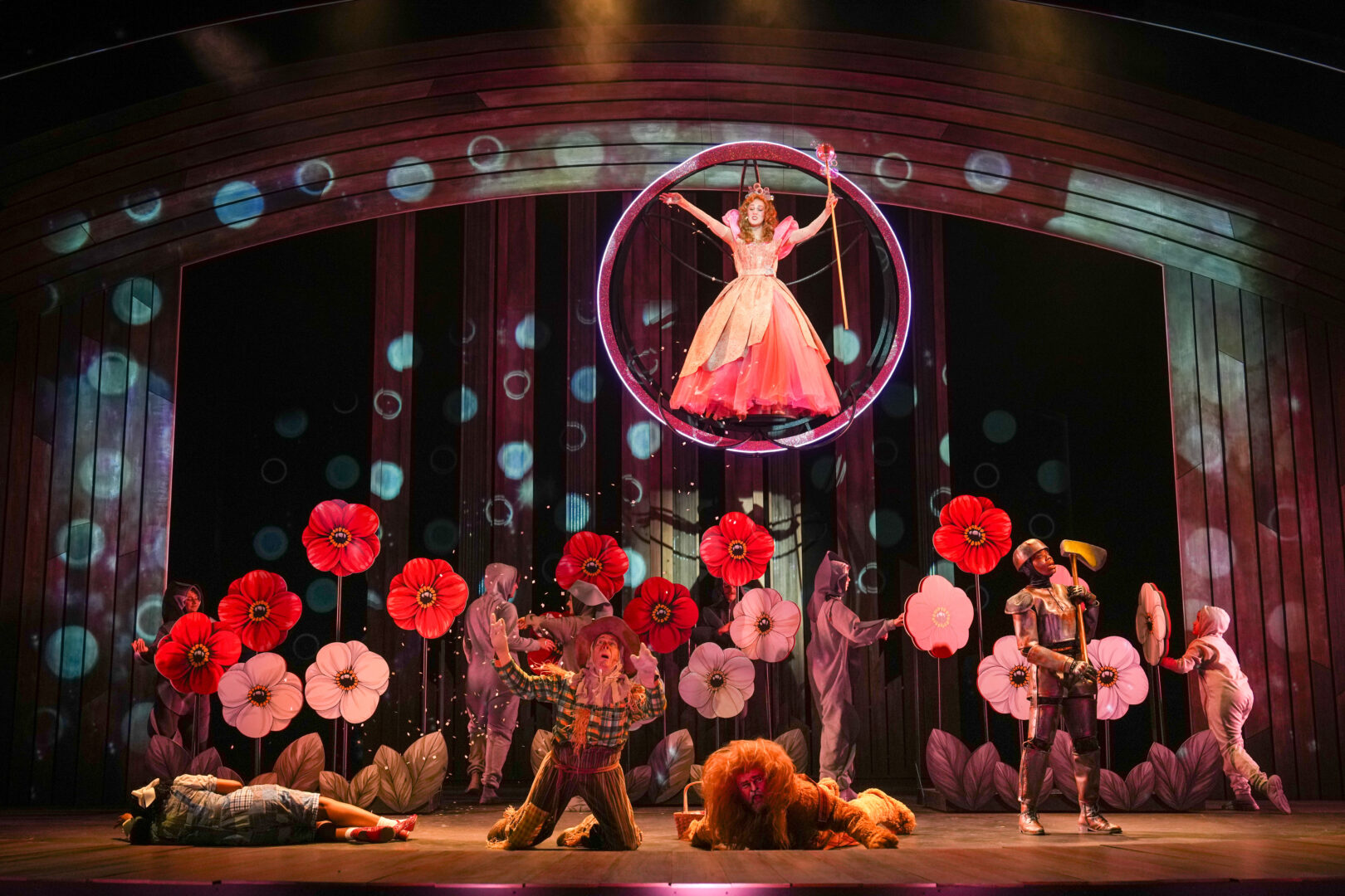 A woman in a pink dress holding a staff floats in a bubble above four people in a field of large red and pink flowers.