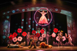 A woman in a pink dress holding a staff floats in a bubble above four people in a field of large red and pink flowers.