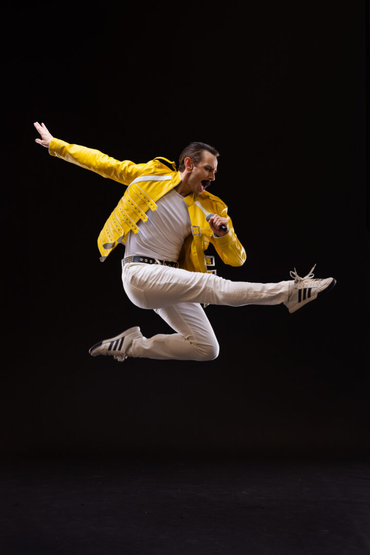 A man in a yellow jacket, white t-shirt, white pants and sneakers is in mid-air, holding a microphone in his left hand, holding his right hand behind him and kicking his right leg forward.