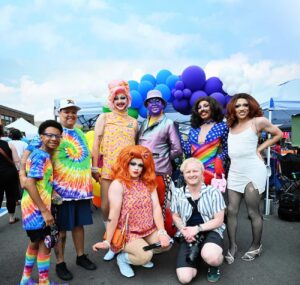 Drag performers and crowd goers pose by a rainbow balloon arch at Anoka Pride festival.