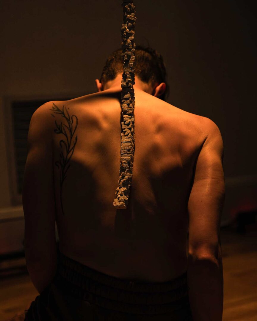 A Body (Un)Becoming solo dancer sits on the ground restrained with her back to the crowd.