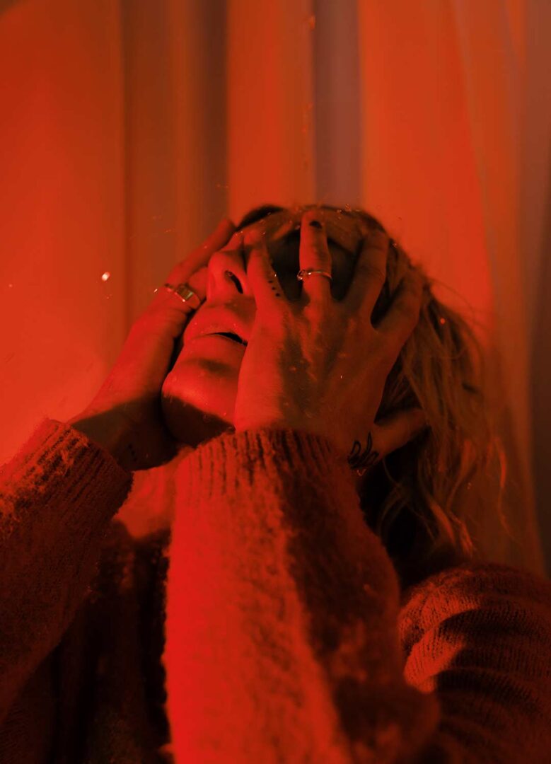 Musical artist POLIÇA in a promotional shot with red lights as she holds her face with her hands.