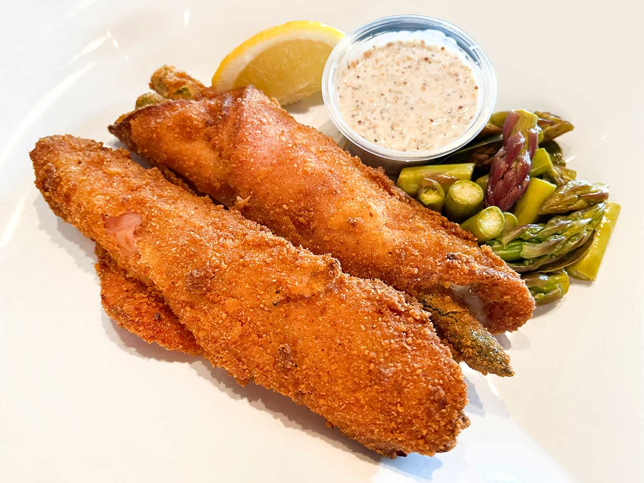 A plate with two Spargel Ham Cordon Bleu with a mustard-mayo sauce and pickled asparagus tips.
