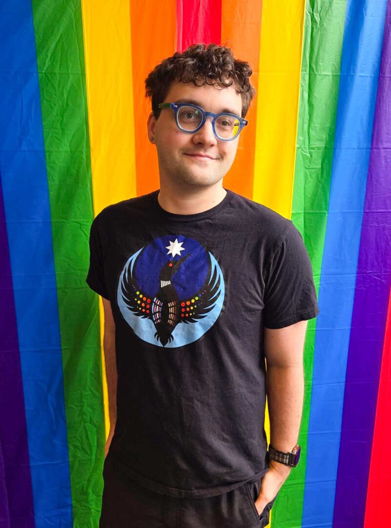 Alexei Dickinson wearing the Pride Loon T-shirt with rainbow flag backdrop.